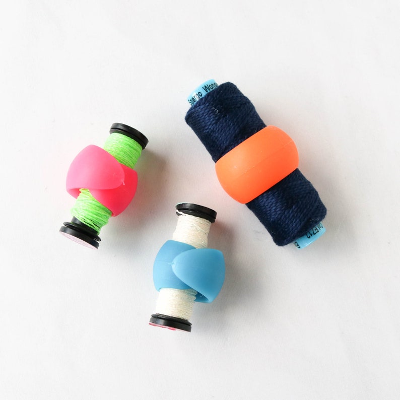 May include: Three spools of thread with colorful silicone thread keepers. The thread colors are green, white, and navy blue. The thread keepers are pink, orange, and blue. The spools are on a white background.