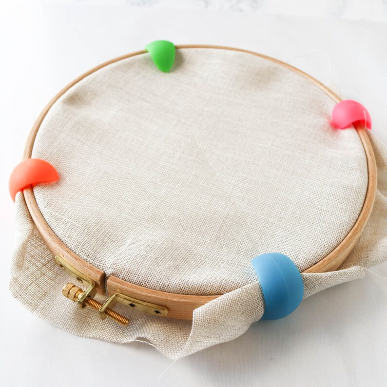 May include: A wooden embroidery hoop with a beige fabric stretched inside. Four colorful silicone clips in orange, pink, green, and blue are attached to the hoop. The hoop is secured with a brass screw.