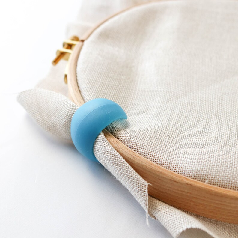 May include: A close-up of an embroidery hoop with a light blue silicone hoop grip. The wooden hoop holds beige fabric. The grip is curved and fits around the wooden hoop.