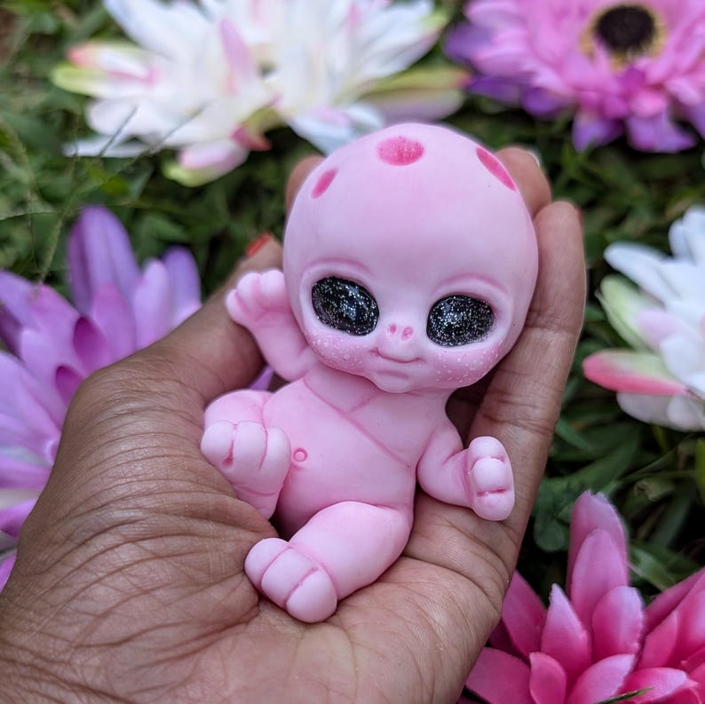 Pode incluir: Uma figura de beb&ecirc; alien&iacute;gena rosa com grandes olhos pretos brilhantes e uma express&atilde;o sorridente. A figura tem tr&ecirc;s manchas vermelhas na cabe&ccedil;a e &eacute; segurada em uma m&atilde;o, cercada por flores rosas e brancas.