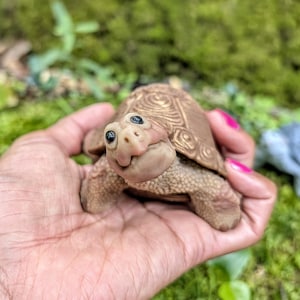 Könnte beinhalten: Eine kleine, hellbraune, skulptierte Schildkrötenfigur mit einem detaillierten, texturierten Panzer, der konzentrische Kreismuster aufweist. Die Schildkröte hat einen glatten, abgerundeten Kopf mit zwei kleinen schwarzen Augen und einem leicht aufwärts gerichteten Mund.