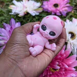 Pode incluir: Uma pequena figura de beb&ecirc; alien&iacute;gena rosa com grandes olhos pretos brilhantes e manchas vermelhas na cabe&ccedil;a. A figura &eacute; segurada em uma m&atilde;o, cercada por flores rosa, roxas e brancas. O beb&ecirc; tem os bra&ccedil;os e as pernas estendidos.