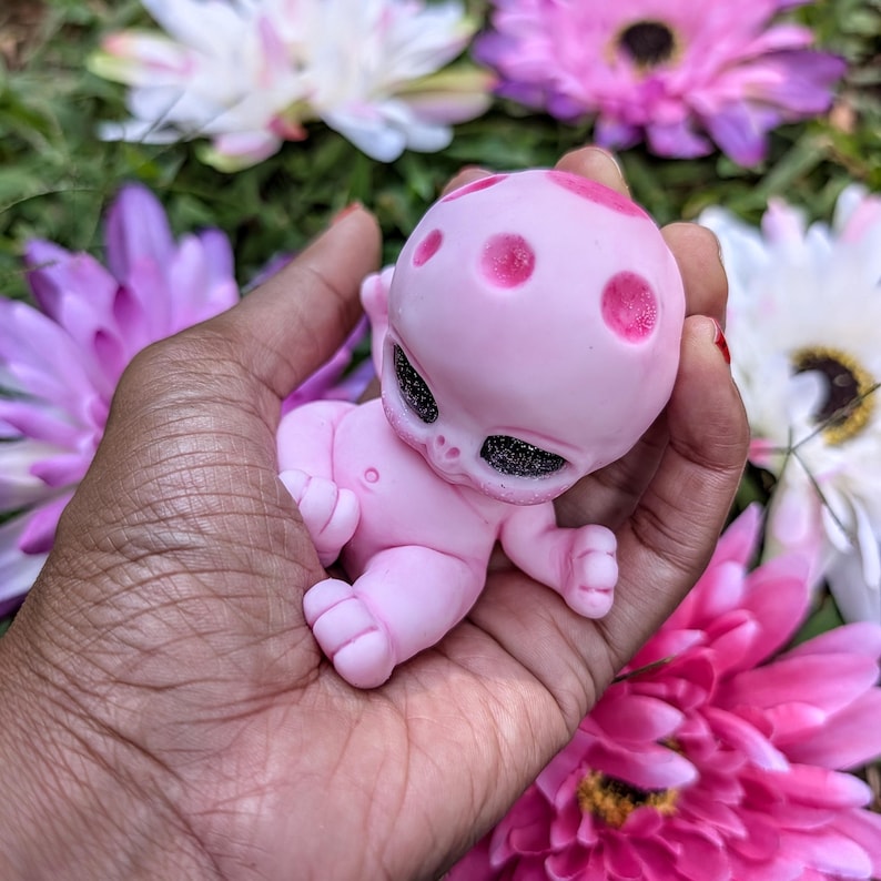 Pode incluir: Uma figura de beb&ecirc; alien&iacute;gena rosa com olhos escuros e manchas rosa na cabe&ccedil;a &eacute; segurada em uma m&atilde;o. A figura &eacute; cercada por flores rosa, roxas e brancas.