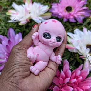 Pode incluir: Uma figura de beb&ecirc; alien&iacute;gena rosa com grandes olhos pretos brilhantes e pequenas manchas vermelhas na cabe&ccedil;a. A figura &eacute; segurada em uma m&atilde;o, cercada por flores rosas, roxas e brancas.