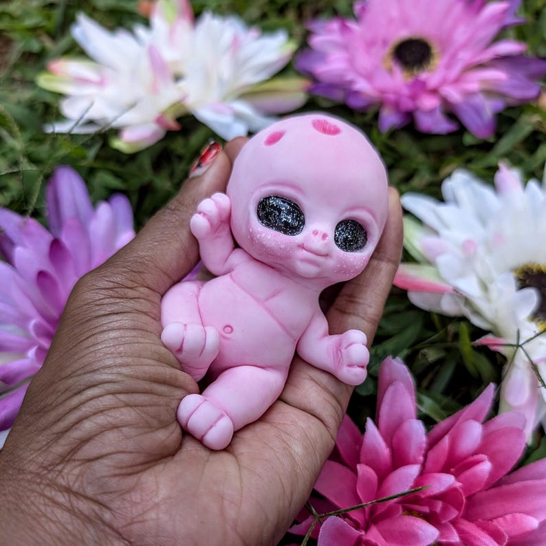 Pode incluir: Uma figura de beb&ecirc; alien&iacute;gena rosa com grandes olhos pretos brilhantes e pequenas manchas vermelhas na cabe&ccedil;a. A figura &eacute; segurada em uma m&atilde;o, cercada por flores rosas, roxas e brancas.