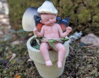 May include: A small porcelain fairy doll with butterfly wings, sleeping on a mushroom in a miniature flower pot. The fairy has a white mushroom cap on its head and is wearing a green leaf sash.