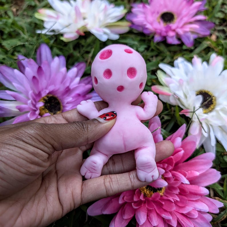 Pode incluir: Uma pequena figura rosa com uma cabe&ccedil;a arredondada e um corpo em forma de beb&eacute;. A cabe&ccedil;a tem v&aacute;rias manchas rosa mais escuras. A figura &eacute; segurada em uma m&atilde;o, rodeada por flores artificiais coloridas.