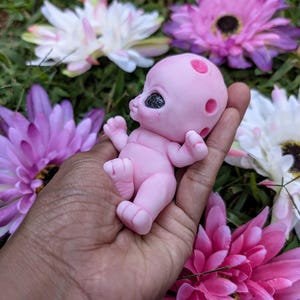 Pode incluir: Uma pequena boneca alien&iacute;gena rosa com olhos escuros e manchas vermelhas na cabe&ccedil;a. A boneca &eacute; segurada em uma m&atilde;o, rodeada por flores rosa e brancas. Os bra&ccedil;os e as pernas da boneca est&atilde;o estendidos.