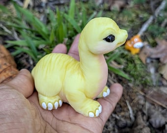 May include: A small, pale yellow dinosaur figurine with a long neck, a smooth texture, and large, dark, glossy eyes. The creature features subtle pink shading on its chest and neck, with white-painted claws on its four feet.