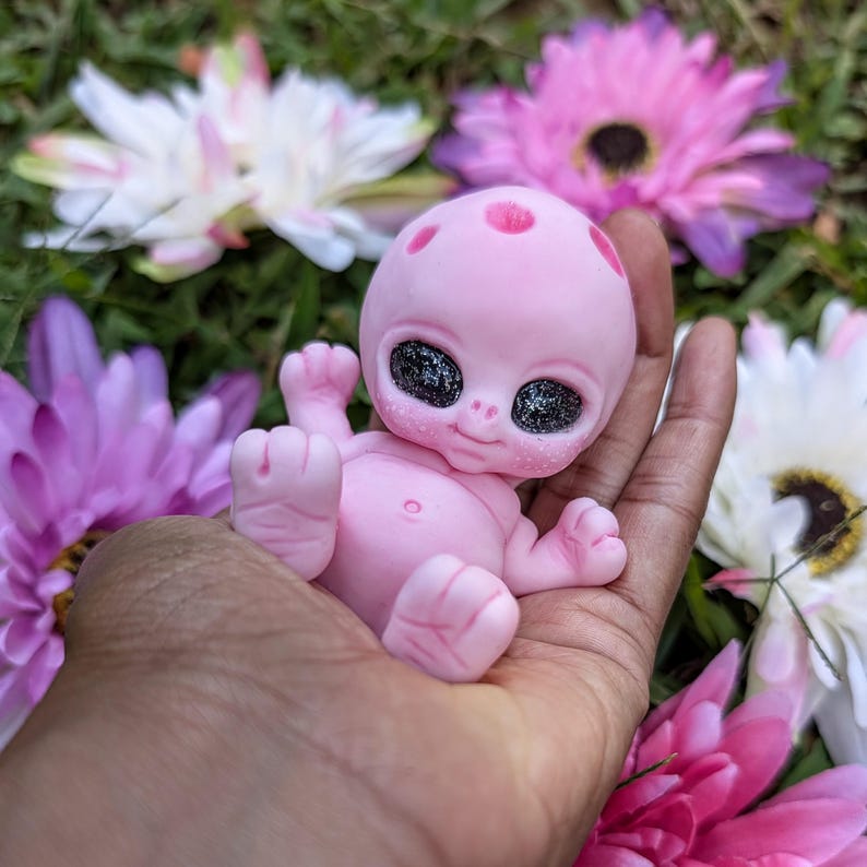 Pode incluir: Uma pequena figura alien&iacute;gena rosa com grandes olhos negros brilhantes e uma express&atilde;o sorridente. A figura &eacute; segurada em uma m&atilde;o, cercada por flores rosa, roxas e brancas. O alien&iacute;gena tem uma cabe&ccedil;a lisa e arredondada com manchas rosa.