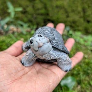 Könnte beinhalten: Eine kleine, modellierte Schildkrötenfigur mit einem strukturierten grauen Panzer mit komplexen Spiralmustern und einem glatten, hellgrauen Kopf mit dunklen, glänzenden Augen. Die Schildkröte hat eine detaillierte, körnige Haut an den Beinen.