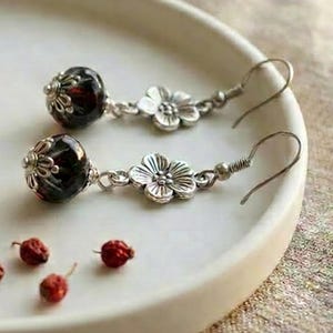 May include: A pair of dangle earrings featuring dark red, spherical beads capped with silver-toned metal filigree, connected to a five-petaled silver-toned flower charm and a curved ear wire. Four small, loose red beads are placed nearby.