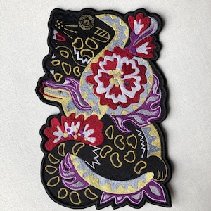 Peut inclure: Patch brodée thermocollante représentant un serpent noir avec des fleurs rouges et blanches. Le serpent est enroulé autour des fleurs et présente un design détaillé avec des accents dorés et violets.