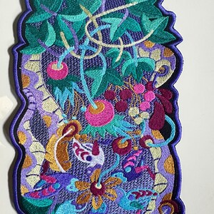 Op de afbeelding: Geborduurde patch met een levendig bloemenontwerp. Voorzien van een paarse achtergrond met gedetailleerde steken in tinten groen, roze, geel en blauw. De patch heeft een geschulpte rand in een donkerdere paarse kleur.