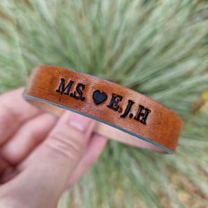Può includere: Un braccialetto in pelle marrone con le iniziali "M.S. ♥ E.J.H" impresse in nero. Il braccialetto ha un interno azzurro chiaro ed è tenuto contro uno sfondo verde sfocato.