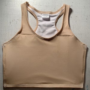 May include: A beige racerback tank top with a white interior lining. The tank top has a smooth, seamless appearance with visible stitching along the edges. The top has a tag that reads "Bodycare Design".