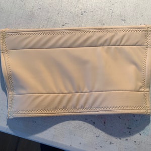 May include: A rectangular, beige-colored padded item with a quilted design. The item has a smooth, matte surface and is stitched around the edges with a matching thread. The item appears to be a protective sleeve or cover.