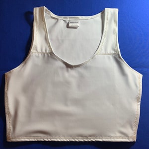 May include: A white sleeveless crop top featuring a scoop neckline and visible stitching along the armholes, neckline, and hem. A small white rectangular tag is attached to the inside of the neckline, displaying the text Medical Care Design.