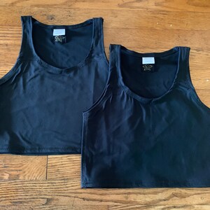 May include: Two black tank tops with a scoop neck and a sleeveless design are displayed on a wooden surface. Each top has a tag with a logo near the neckline. The tops are made of a smooth, solid fabric.