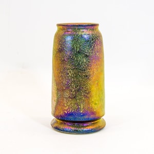 Könnte beinhalten: Eine zylindrische, schillernde Vase mit strukturierter Oberfläche. Die Vase hat einen breiten Sockel und eine etwas breitere Oberseite. Die Farben wechseln von Gold über Grün zu Blau und erzeugen einen schimmernden Effekt. Die Vase ist etwa 18 cm hoch.