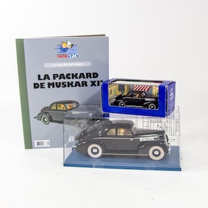 Op de afbeelding: Een zwart-wit stripboek met de titel "La Packard de Muskar XI" met een zwarte auto op de omslag. Het stripboek staat naast een transparante vitrine met een zwarte speelgoedauto erin. De vitrine staat op een blauwe doos met een zwarte auto op de omslag.
