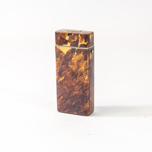 Puede incluir: Una pequeña caja rectangular hecha de carey con un borde dorado. La caja tiene un patrón moteado marrón y amarillo.