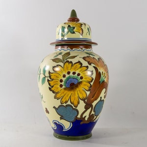 Może przedstawiać: Ceramiczny wazon z pokrywką, ozdobiony kwiatowym wzorem w odcieniach żółtego, niebieskiego, zielonego i brązowego. Wazon ma kremowy kolor tła i niebieską podstawę.