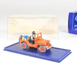 May include: A red toy jeep with a black driver figure and a blue and white box. The jeep has a black roof and black wheels. The box has the words 'The Toys of Our Time' printed on the bottom.