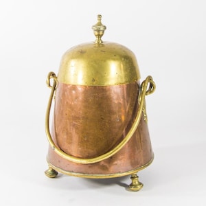 Könnte beinhalten: Ein antiker Kohlenkübel aus Kupfer und Messing mit einem Messingdeckel und -griff. Der Kübel hat drei Beine und einen abgerundeten Korpus.