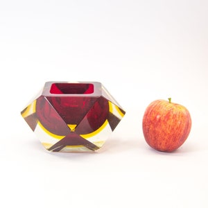 Puede incluir: Un cenicero geométrico de vidrio rojo y amarillo con un diseño facetado. El cenicero está sobre una superficie blanca junto a una manzana roja.
