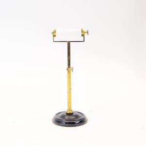 Könnte beinhalten: Ein Toilettenpapierhalter im Vintage-Stil. Der Ständer hat einen schwarzen, kreisförmigen Sockel und eine verstellbare Stange in Messingfarbe. Die Toilettenpapierrolle wird von einer messingfarbenen Stange gehalten. Der Halter ist für den Boden konzipiert.