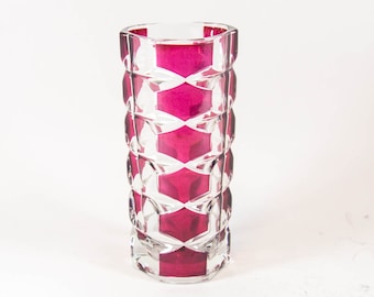 May include: A cylindrical glass vase featuring a deep magenta interior layer encased in clear glass. The exterior surface is decorated with a repeating geometric pattern of vertical facets and horizontal cut-out shapes.