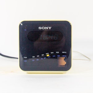 May include: A vintage Sony digital clock radio with a black face and a yellow trim. The clock radio has a digital display showing the time and a dial for tuning radio stations. The radio is plugged in with a black cord.