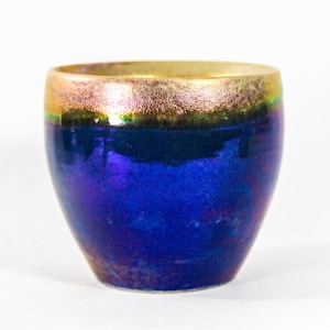 Könnte beinhalten: Ein kleiner, runder, blauer Keramiktopf mit goldfarbenem Rand. Der Topf hat eine irisierende Glasur, die im Licht schimmert.