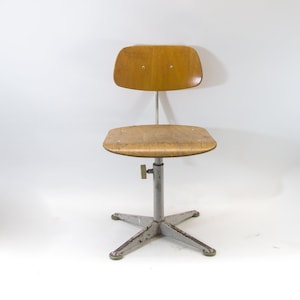 Puede incluir: Una silla giratoria vintage de madera y metal con base en forma de estrella. La silla tiene un asiento y un respaldo de madera clara.