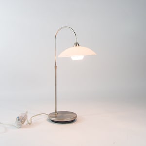 Può includere: Una lampada da tavolo argentata con un paralume bianco e un braccio curvo. La lampada ha una base rotonda e un cavo con una spina.