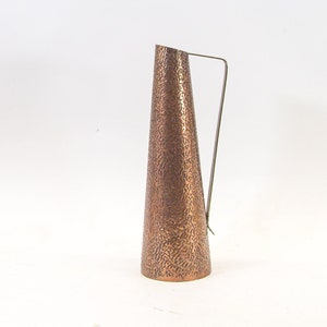 May include: A hammered copper vase with a slender, conical shape and a thin metal handle. The vase has a textured surface and a warm, metallic color.