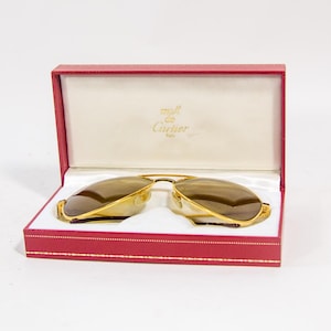Può includere: Occhiali da sole aviator con montatura dorata e lenti marroni in una scatola rossa aperta. La scatola mostra un interno color crema con il logo Cartier. Gli occhiali hanno un design classico.