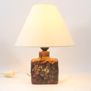 May include: A ceramic table lamp with a cream-colored lampshade. The lamp base is rectangular with a textured, mottled brown and orange glaze. A white cord with an on/off switch extends from the base.