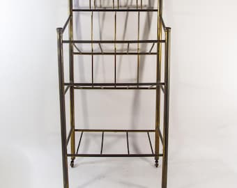 May include: An antique metal magazine rack with a dark bronze finish and gold accents. The rack has a rectangular shape with multiple shelves and sits on small caster wheels. The design is vertical and features a grid-like pattern.