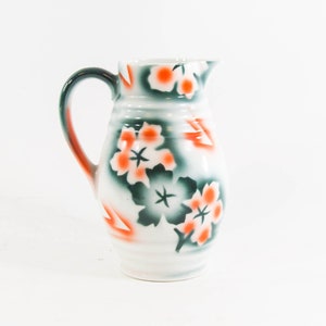Op de afbeelding: Een witte keramische kan met een groen en oranje bloemenmotief. De kan heeft een handvat en een rond lichaam.