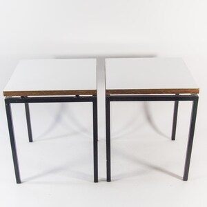 Op de afbeelding: Een paar vierkante bijzettafels met witte tafelbladen en donkere houten randen, ondersteund door dunne zwarte metalen frames. Elke tafel heeft vier rechte poten en een eenvoudig ontwerp dat een vlak oppervlak biedt voor gebruik.