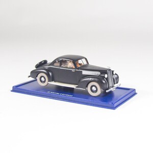 Op de afbeelding: Zwarte model auto van gegoten metaal op een blauwe display basis. De auto heeft een wit interieur en witte banden. De basis heeft de tekst "1:24  DETROIT  2-DOOR  SEDAN".