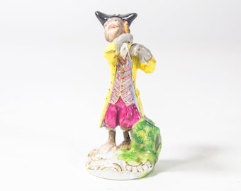May include: A porcelain figurine of a monkey dressed in a yellow jacket, red pants, and a black hat. The monkey is holding a musical instrument and standing on a green base.