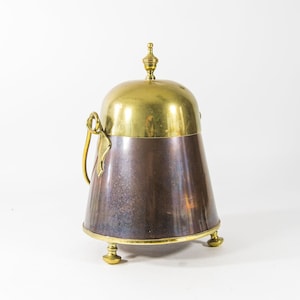 Könnte beinhalten: Eine Teedose aus Messing und Kupfer mit einem gewölbten Deckel und drei Beinen. Der Deckel hat einen Knauf oben.