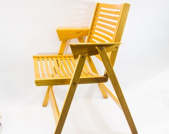 May include: A light wood folding chair with a slatted back and seat. The chair has a small armrest on one side and a simple, sturdy design.