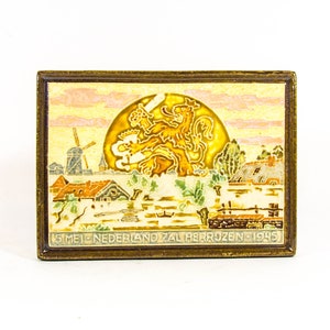 Könnte beinhalten: Eine goldfarbene Metallstifte mit einem bunten Emaille-Design. Das Design zeigt einen Löwen, eine Windmühle und einen Sonnenuntergang mit dem Text "15 MEI NEDERLAND ZAL HERRIJZEN 1945".