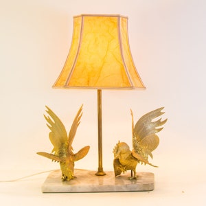May include: A table lamp with a yellow lampshade and two gold rooster figurines on a white marble base.