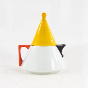 Può includere: Una teiera in ceramica bianca con coperchio conico giallo e piccolo pomello giallo. Il manico è arancione brillante e il beccuccio è nero. La teiera ha un design moderno e geometrico.