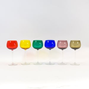 Op de afbeelding: Zes glazen met steel in verschillende kleuren: rood, geel, groen, blauw, roze en bruin. De glazen zijn in een rij op een wit oppervlak gerangschikt.